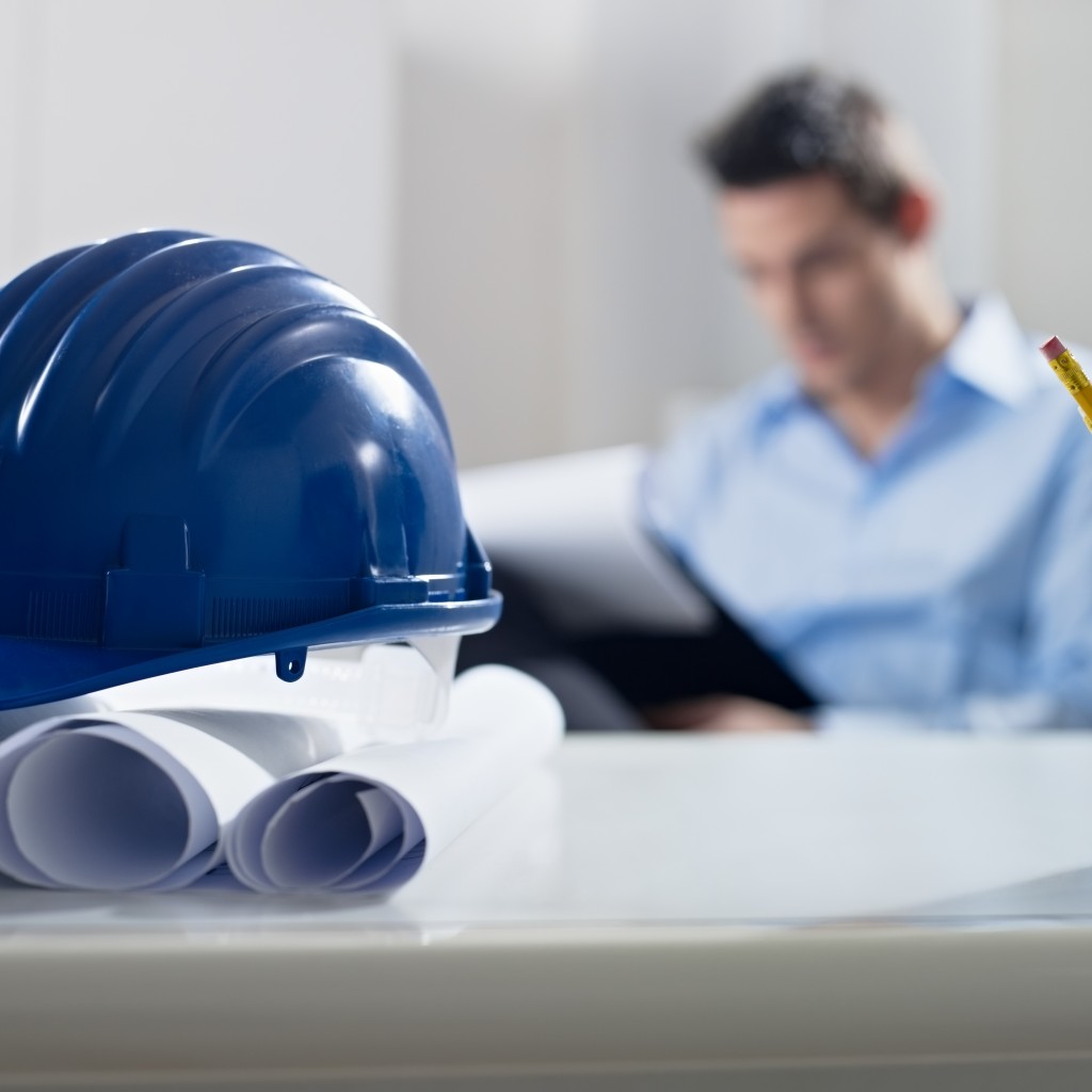 adult caucasian male architect examining documents. Focus on blueprints and hardhat in foreground. Horizontal shape, front view